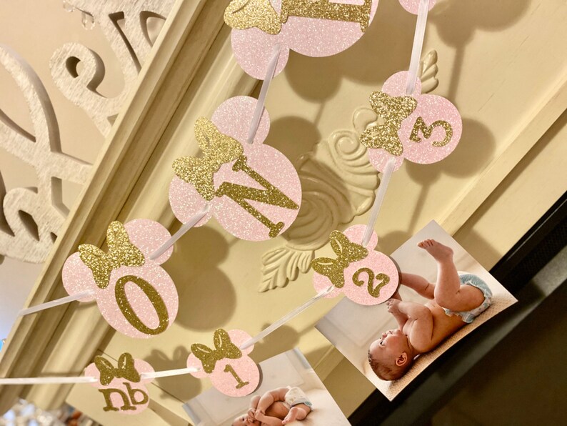 May include: Pink and gold glitter Minnie Mouse themed banner with the numbers 0-3 and the letters "nb" for a first birthday party.