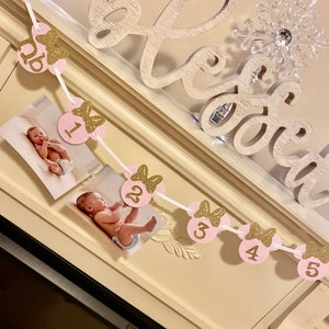Op de afbeelding: Een roze en gouden glitterbanner met Minnie Mouse-thema met de nummers 1 tot en met 5 en een strik op elke cirkel. De banner is aan een wit lint geregen en heeft foto's van een babymeisje in elke cirkel.