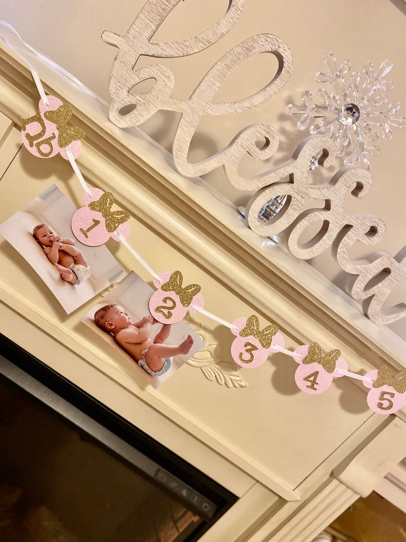 Op de afbeelding: Een roze en gouden glitterbanner met Minnie Mouse-thema met de nummers 1 tot en met 5 en een strik op elke cirkel. De banner is aan een wit lint geregen en heeft foto's van een babymeisje in elke cirkel.