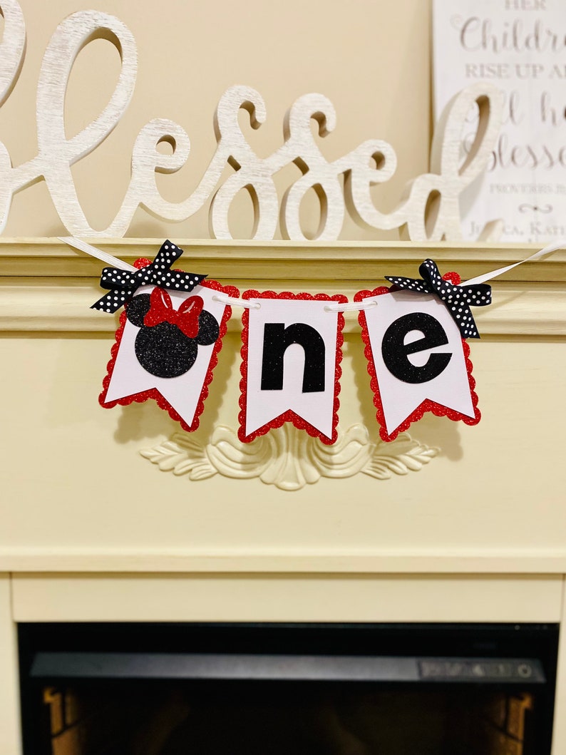 May include: A decorative banner featuring three white pennants with scalloped red glitter edges. The first pennant displays a black glitter mouse silhouette with a red bow, while the others show the black letters n and e. Black and white polka dot bows are attached to the top corners.
