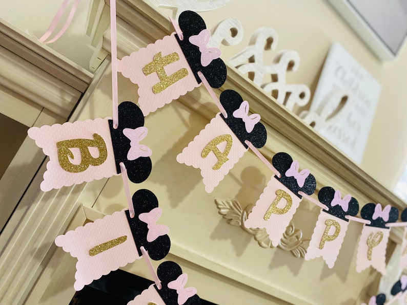 May include: Pink and black banner with gold glitter letters spelling "Happy Birthday". Each letter is attached to a black Mickey Mouse ear with a pink bow.