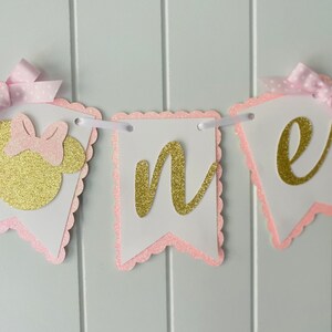 Puede incluir: Bandera de brillo rosa y dorado con la palabra "uno" y una cabeza de Minnie Mouse. La bandera es perfecta para una fiesta de primer cumplea&ntilde;os.