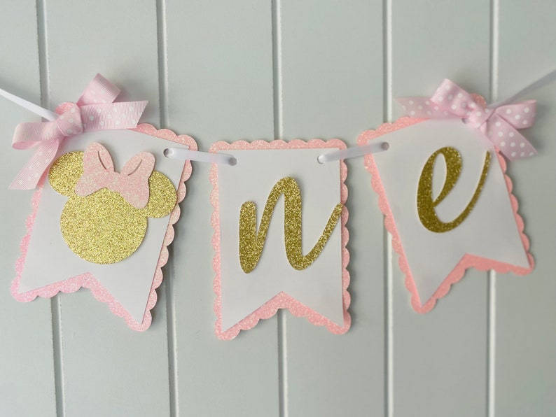 Puede incluir: Bandera de brillo rosa y dorado con la palabra "uno" y una cabeza de Minnie Mouse. La bandera es perfecta para una fiesta de primer cumplea&ntilde;os.