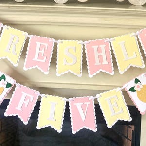 May include: A two-tiered decorative banner featuring scalloped cardstock pennants in alternating pastel pink and yellow. The top row spells FRESHLY and the bottom row spells FIVE in white letters. Two glittery yellow lemon cutouts flank the text.