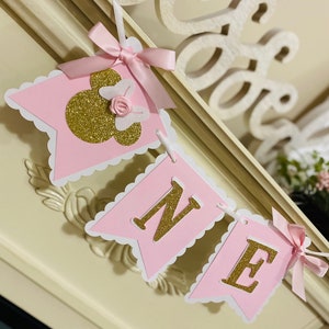 Puede incluir: Bandera rosa y dorada con la palabra "ONE" y una cabeza de Minnie Mouse con un lazo rosa. La bandera est&aacute; decorada con bordes festoneados blancos.