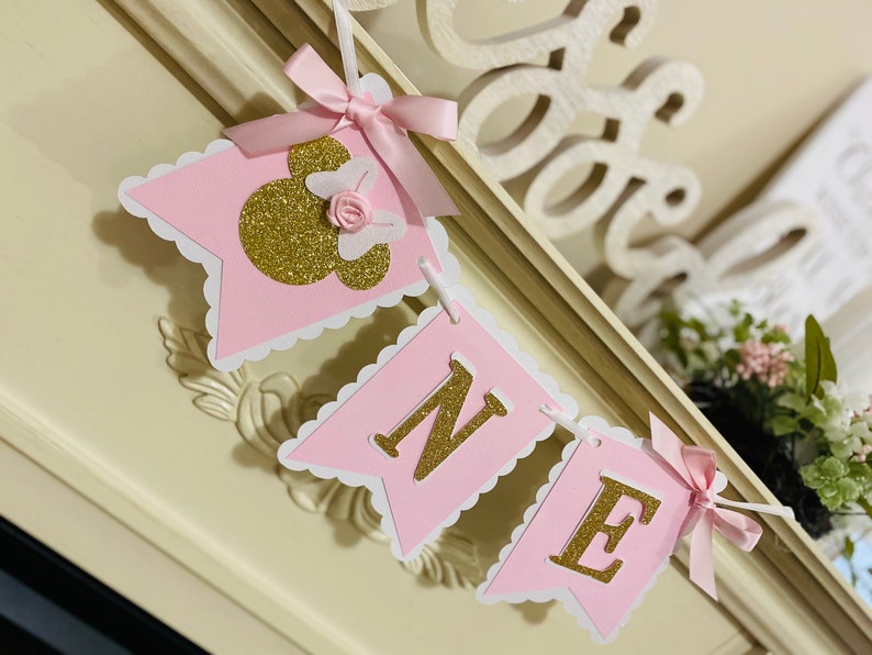 Puede incluir: Bandera rosa y dorada con la palabra "ONE" y una cabeza de Minnie Mouse con un lazo rosa. La bandera est&aacute; decorada con bordes festoneados blancos.
