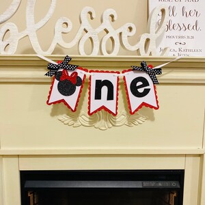 May include: A decorative banner featuring three white pennants with red scalloped edges, spelling the word one. The first pennant displays a black glittery mouse silhouette with a red bow, while the others feature black glittery letters.