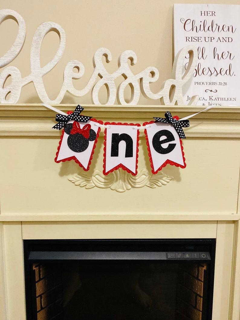 May include: A decorative banner featuring three white pennants with red scalloped edges, spelling the word one. The first pennant displays a black glittery mouse silhouette with a red bow, while the others feature black glittery letters.