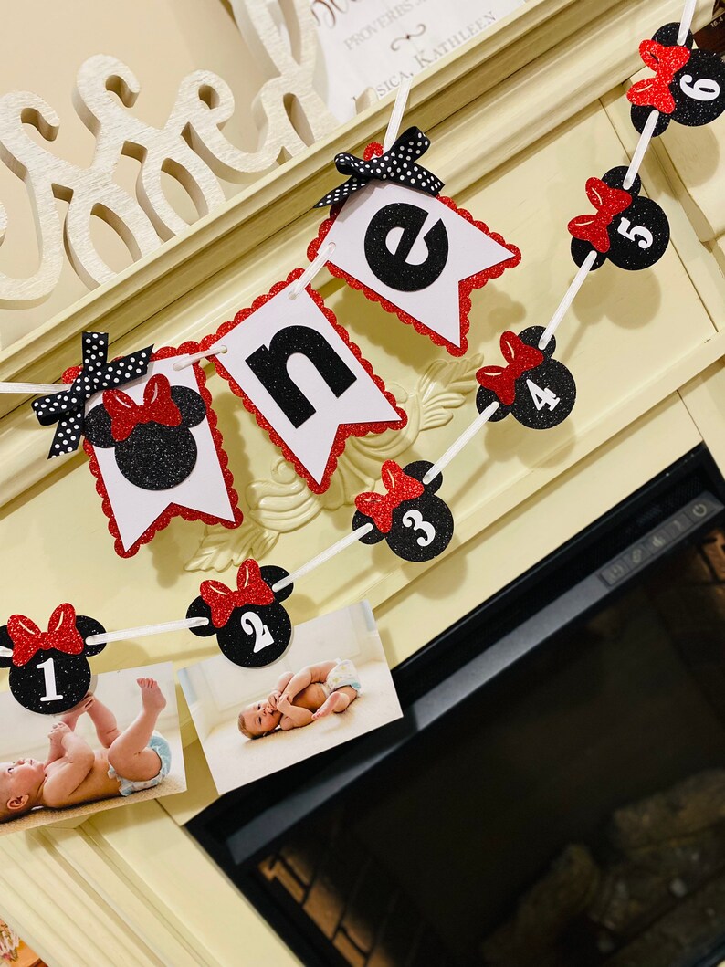 May include: A red and black Minnie Mouse themed banner with the word "one" and numbers 1 through 6. The banner is strung with white string and has red bows with black polka dots. There are photos of a baby in the bottom of the image.
