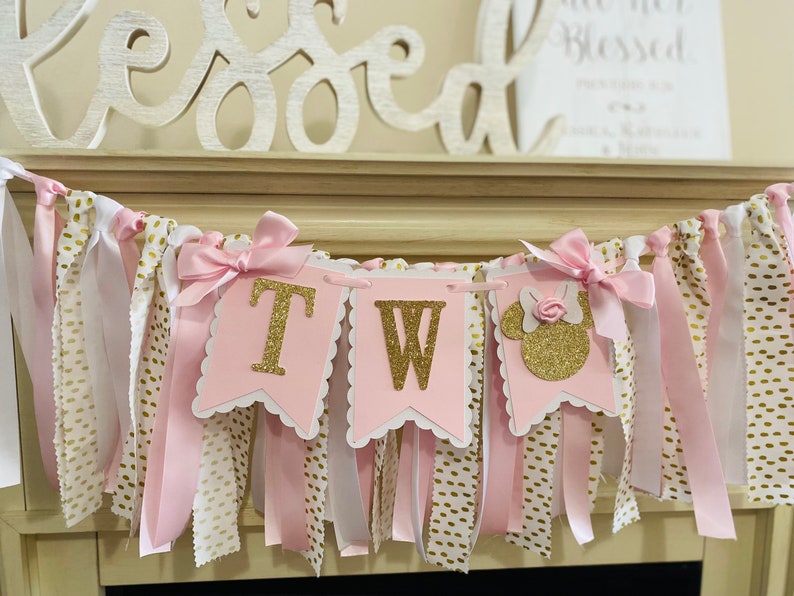 May include: A pink and gold banner with the letters "TW" and a Minnie Mouse silhouette. The banner is decorated with pink ribbons, white fabric with gold polka dots, and pink bows. The word "Blessed" is visible in the background.