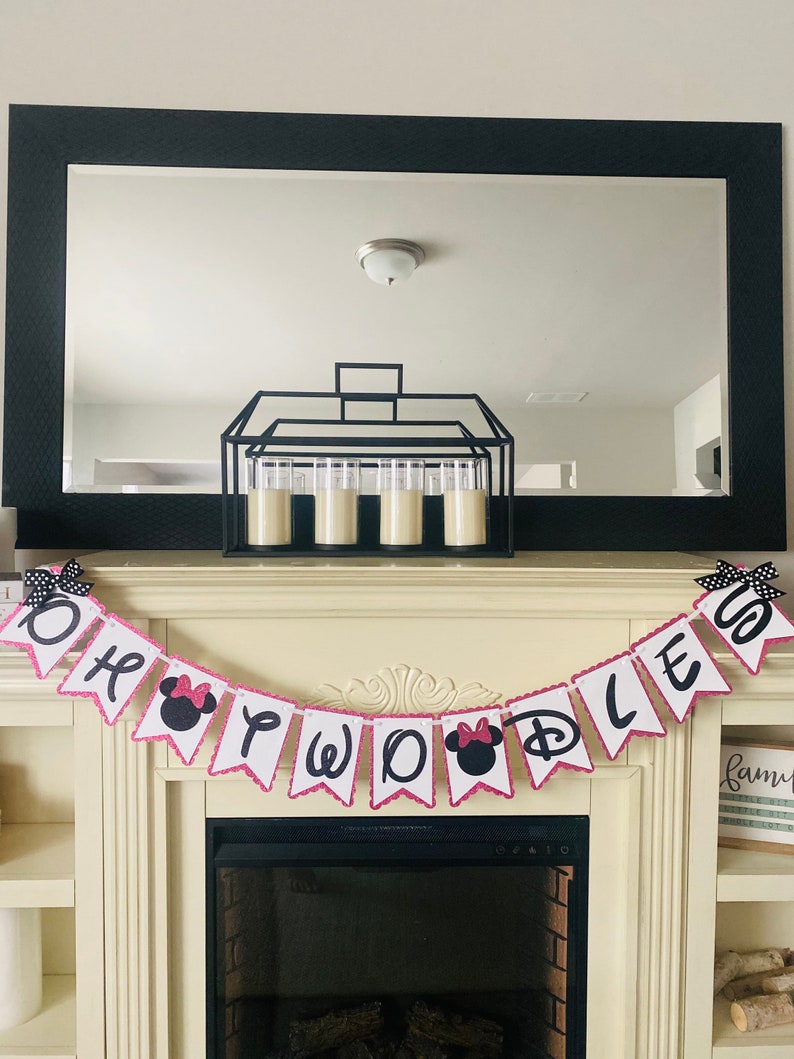 May include: A white and pink banner with black letters that reads "Oh Two D" with a pink Minnie Mouse head on each of the "O"s. The banner is hanging above a fireplace.
