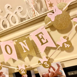 Puede incluir: Bandera de brillo rosa y dorado con la palabra "ONE" y orejas de Minnie Mouse con los n&uacute;meros del 1 al 5. La bandera est&aacute; enhebrada en una cuerda blanca y est&aacute; colgada de una repisa blanca.