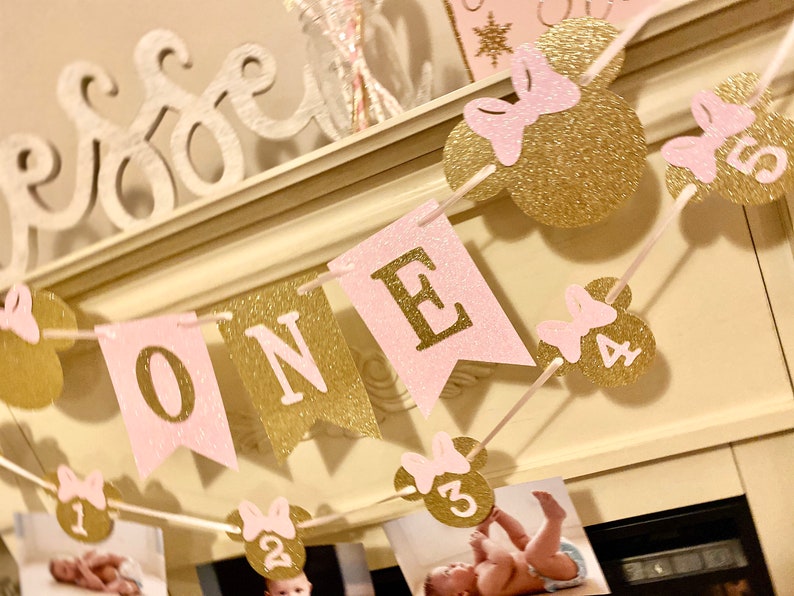 Puede incluir: Bandera de brillo rosa y dorado con la palabra "ONE" y orejas de Minnie Mouse con los n&uacute;meros del 1 al 5. La bandera est&aacute; enhebrada en una cuerda blanca y est&aacute; colgada de una repisa blanca.