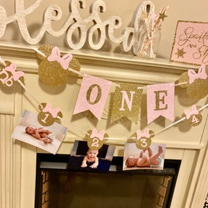 Puede incluir: Una pancarta de brillo rosa y dorado con la palabra "ONE" y orejas de Mickey Mouse con lazos rosas. La pancarta est&aacute; decorada con fotos de una ni&ntilde;a peque&ntilde;a a diferentes edades. La pancarta est&aacute; colgada sobre una chimenea.