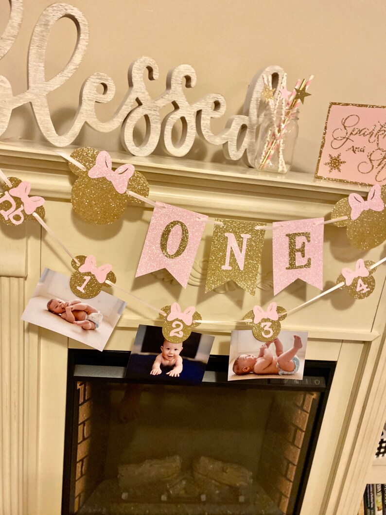 Puede incluir: Una pancarta de brillo rosa y dorado con la palabra "ONE" y orejas de Mickey Mouse con lazos rosas. La pancarta est&aacute; decorada con fotos de una ni&ntilde;a peque&ntilde;a a diferentes edades. La pancarta est&aacute; colgada sobre una chimenea.