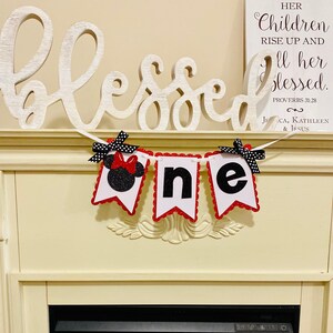 May include: A decorative banner featuring three white pennants with red scalloped edges, spelling out the word one. The first pennant displays a black glittery mouse silhouette with a red bow, while the others show black glittery letters.