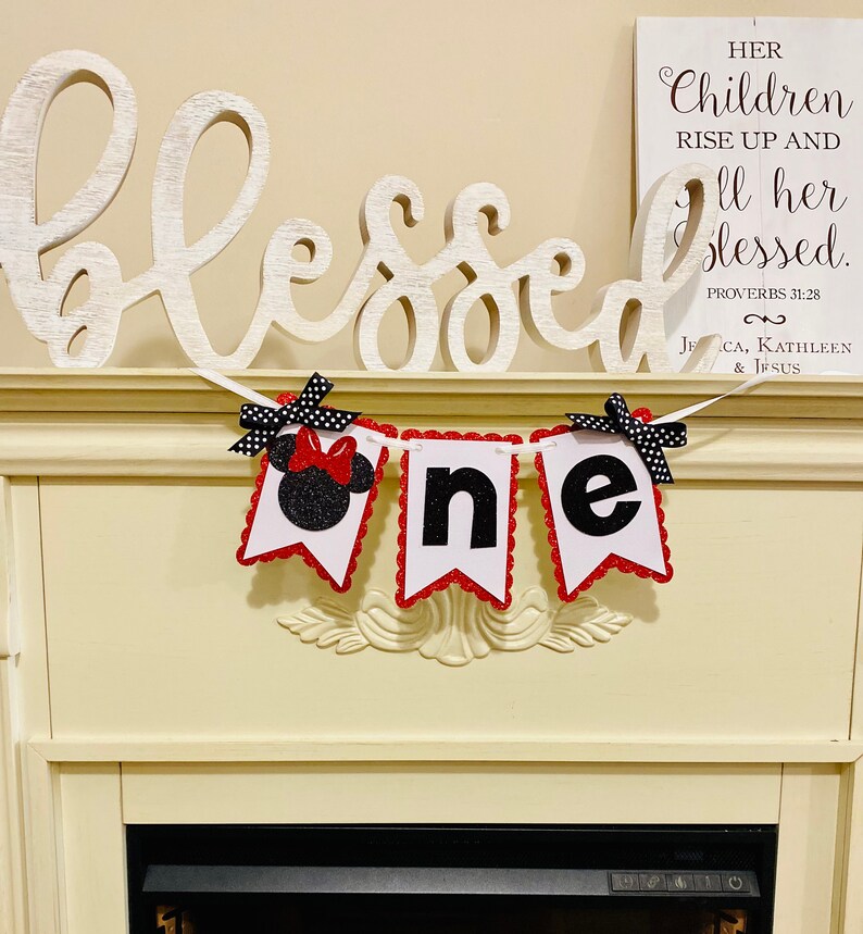 May include: A decorative banner featuring three white pennants with red scalloped edges, spelling out the word one. The first pennant displays a black glittery mouse silhouette with a red bow, while the others show black glittery letters.