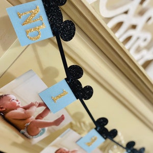 Pode incluir: Uma faixa azul e preta com letras douradas brilhantes que soletram "Um" e orelhas do Mickey Mouse. A faixa &eacute; decorada com fotos de um beb&ecirc;.