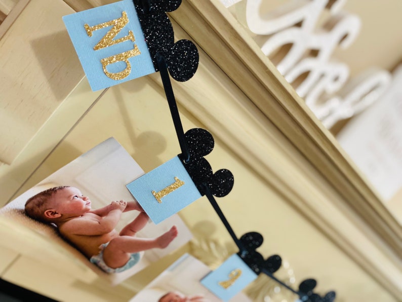 Pode incluir: Uma faixa azul e preta com letras douradas brilhantes que soletram "Um" e orelhas do Mickey Mouse. A faixa &eacute; decorada com fotos de um beb&ecirc;.