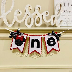 May include: A decorative banner spelling the word one, featuring three white pennants with scalloped red glitter edges. The first pennant displays a black glitter mouse silhouette with a red glitter bow, while the others show black letters.