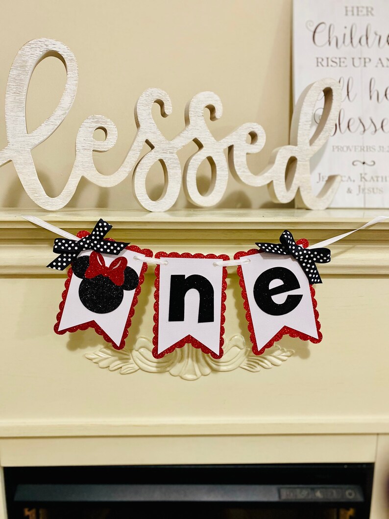 May include: A decorative banner spelling the word one, featuring three white pennants with scalloped red glitter edges. The first pennant displays a black glitter mouse silhouette with a red glitter bow, while the others show black letters.