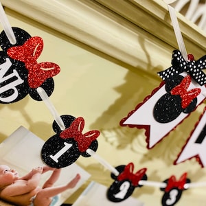 May include: A black and red Minnie Mouse themed banner with the letters "ND" and the numbers "1" through "9" on each of the individual flags. The banner is strung together with white string and has red glitter bows on each flag.
