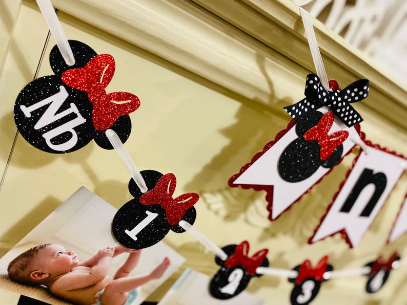May include: A black and red Minnie Mouse themed banner with the letters "ND" and the numbers "1" through "9" on each of the individual flags. The banner is strung together with white string and has red glitter bows on each flag.