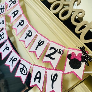 May include: Pink and white banner with black glitter letters that reads "Happy 2nd Birthday". The banner has a pink scalloped edge and a black and white Minnie Mouse head at the end.