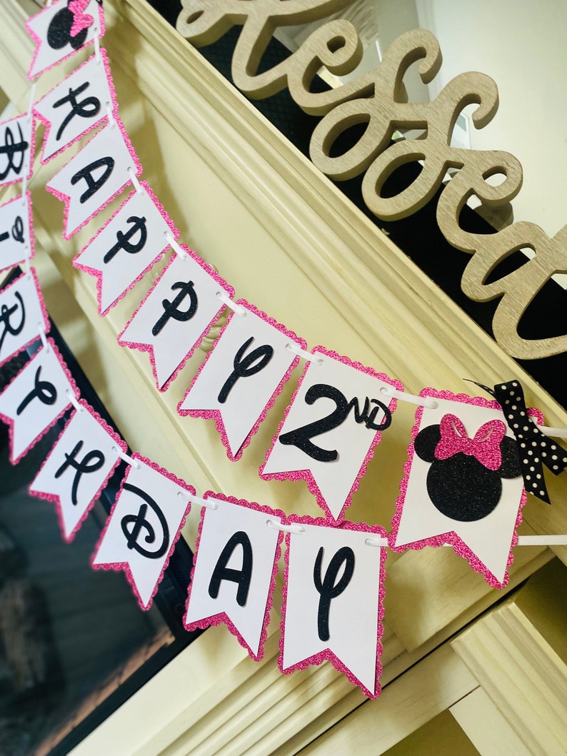 May include: Pink and white banner with black glitter letters that reads "Happy 2nd Birthday". The banner has a pink scalloped edge and a black and white Minnie Mouse head at the end.