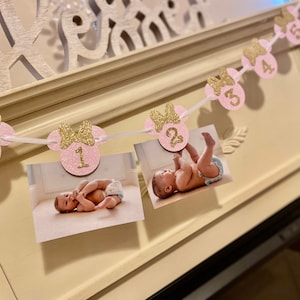 Op de afbeelding: Roze en gouden glitter Minnie Mouse fotobanner met de nummers 1 tot en met 5. Elk nummer staat op een aparte roze cirkel met een gouden strik. De banner is aan een wit lint geregen en heeft foto's van een babymeisje bij elk nummer.
