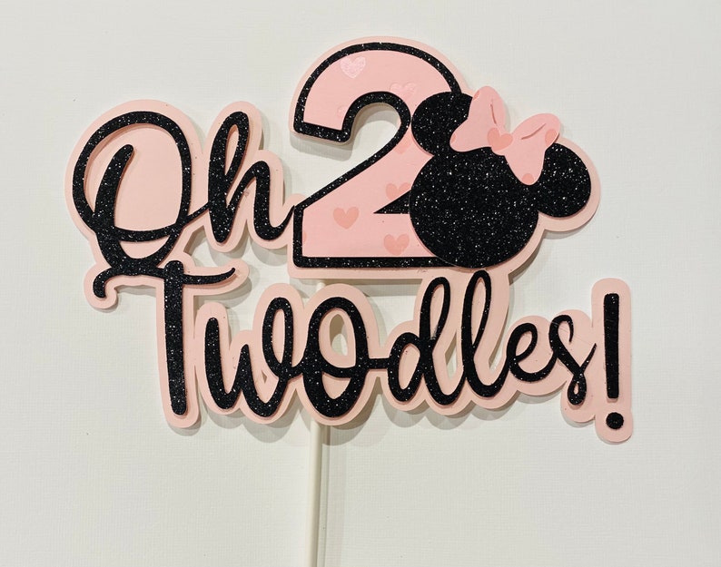 May include: Pink and black cake topper with the text "Oh Two-dles!" and a number 2 with pink hearts. A black silhouette of a mouse head with a pink bow is next to the number 2.