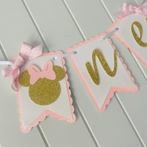 Puede incluir: Bandera de brillo rosa y dorado con la palabra "uno" y una cabeza de Minnie Mouse con un lazo rosa.