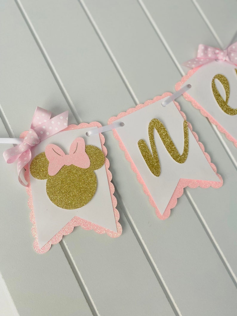 Puede incluir: Bandera de brillo rosa y dorado con la palabra "uno" y una cabeza de Minnie Mouse con un lazo rosa.