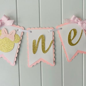 Puede incluir: Pancarta de "uno" con purpurina rosa y dorada con una cabeza de Minnie Mouse de purpurina rosa y dorada. La pancarta es perfecta para una fiesta de primer cumplea&ntilde;os.