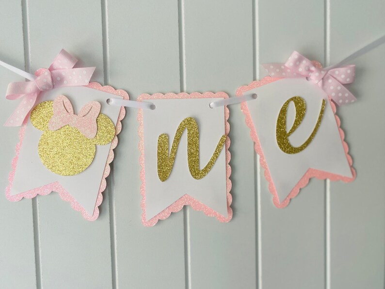 Puede incluir: Pancarta de "uno" con purpurina rosa y dorada con una cabeza de Minnie Mouse de purpurina rosa y dorada. La pancarta es perfecta para una fiesta de primer cumplea&ntilde;os.