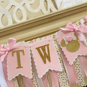 May include: Pink and gold banner with the letters "TW" in gold glitter, along with a Minnie Mouse silhouette. The banner has pink and white ribbons, and is decorated with pink bows. The background includes a wooden "Blessed" sign.