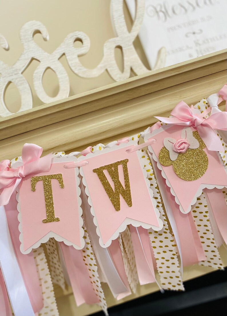 May include: Pink and gold banner with the letters "TW" in gold glitter, along with a Minnie Mouse silhouette. The banner has pink and white ribbons, and is decorated with pink bows. The background includes a wooden "Blessed" sign.