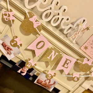 Puede incluir: Una pancarta de brillo rosa y dorado con la palabra "ONE" y orejas de Mickey Mouse con lazos rosas. La pancarta est&aacute; enhebrada con fotos de una ni&ntilde;a peque&ntilde;a a diferentes edades.