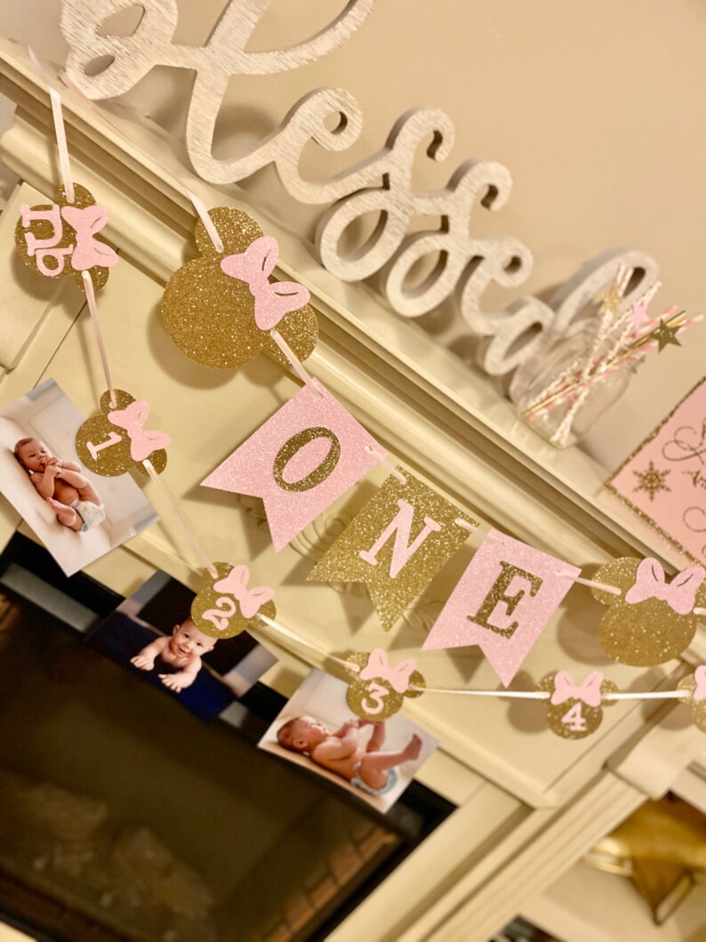 Puede incluir: Una pancarta de brillo rosa y dorado con la palabra "ONE" y orejas de Mickey Mouse con lazos rosas. La pancarta est&aacute; enhebrada con fotos de una ni&ntilde;a peque&ntilde;a a diferentes edades.