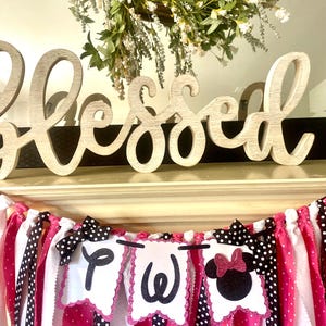 May include: A decorative banner with pink, white, and black polka dot ribbons and pennants. The pennants spell out a word with black letters and feature a Minnie Mouse design. A wooden "Blessed" sign sits above the banner, with a floral wreath in the background.