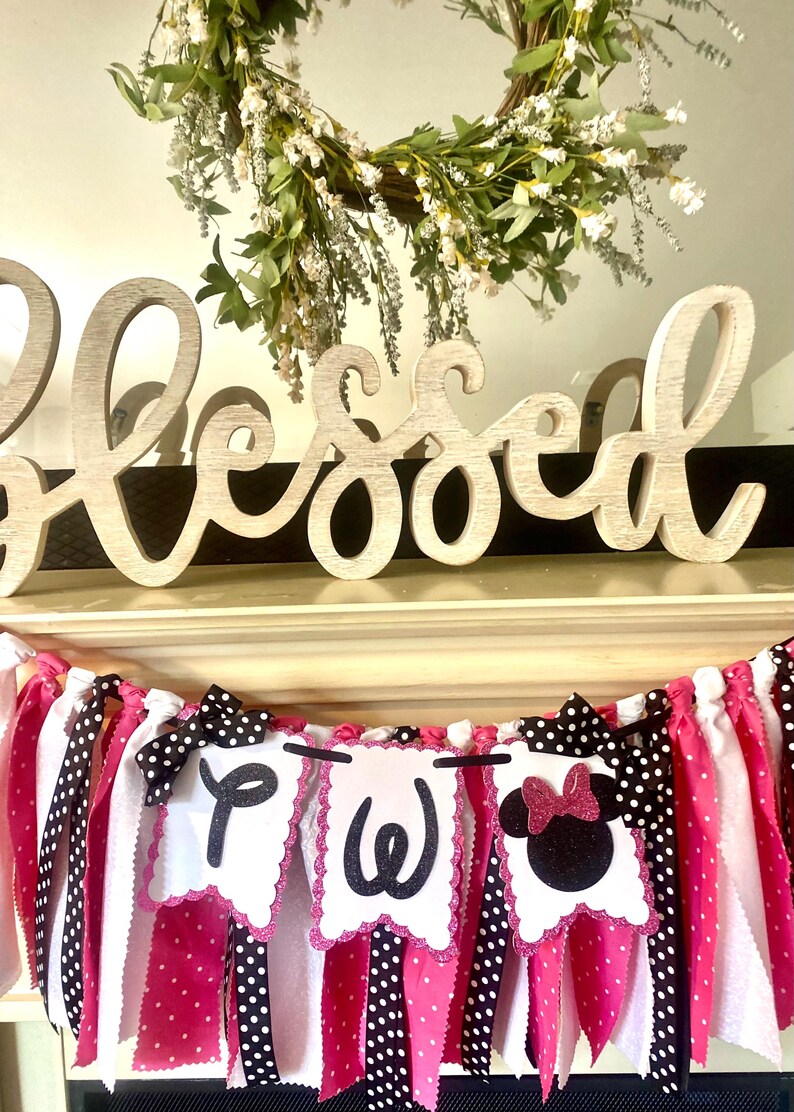 May include: A decorative banner with pink, white, and black polka dot ribbons and pennants. The pennants spell out a word with black letters and feature a Minnie Mouse design. A wooden "Blessed" sign sits above the banner, with a floral wreath in the background.