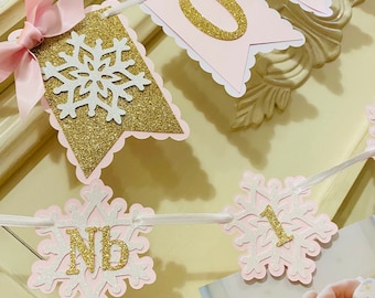 May include: A pink and gold banner with snowflake and number cutouts. The banner includes the letters "NB" and the number "1". A pink bow is attached to the banner. A photo of a baby is in the background.