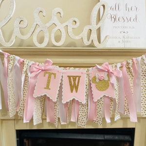 May include: A pink and gold "TWO" banner with a Minnie Mouse silhouette, perfect for a second birthday. The banner has pink, white, and gold ribbons and bows. A wooden sign with the word "Blessed" and a Bible verse is in the background.