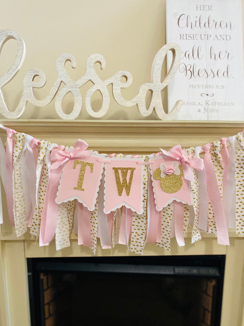 May include: A pink and gold "TWO" banner with a Minnie Mouse silhouette, perfect for a second birthday. The banner has pink, white, and gold ribbons and bows. A wooden sign with the word "Blessed" and a Bible verse is in the background.