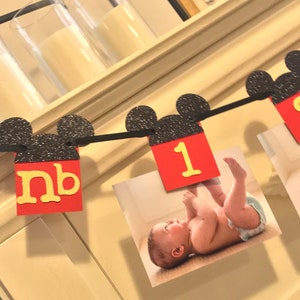 May include: A black and red Mickey Mouse themed banner with the number "1" and the letters "nb" in yellow. The banner is decorated with black glitter and has photos of a baby attached to it.