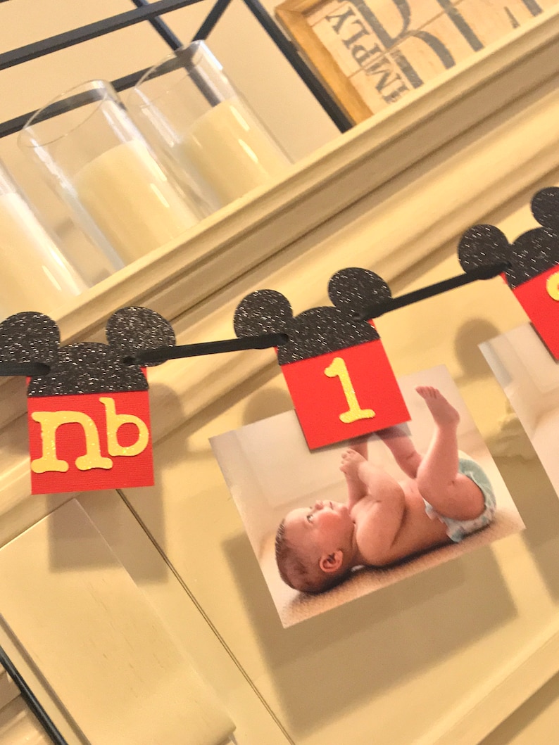May include: A black and red Mickey Mouse themed banner with the number "1" and the letters "nb" in yellow. The banner is decorated with black glitter and has photos of a baby attached to it.