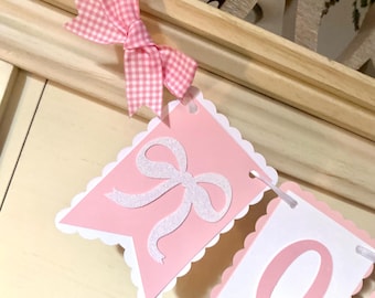 May include: Pink and white banner with a scalloped edge. The banner has a white bow with glitter on a pink background and a white number 0 on a pink background.