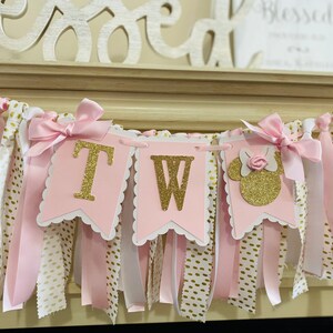 May include: A pink and gold banner with the letters "TW" and a Minnie Mouse silhouette. The banner is decorated with pink ribbons, white ribbons with gold polka dots, and gold glitter accents. A decorative piece that says "Blessed" is in the background.