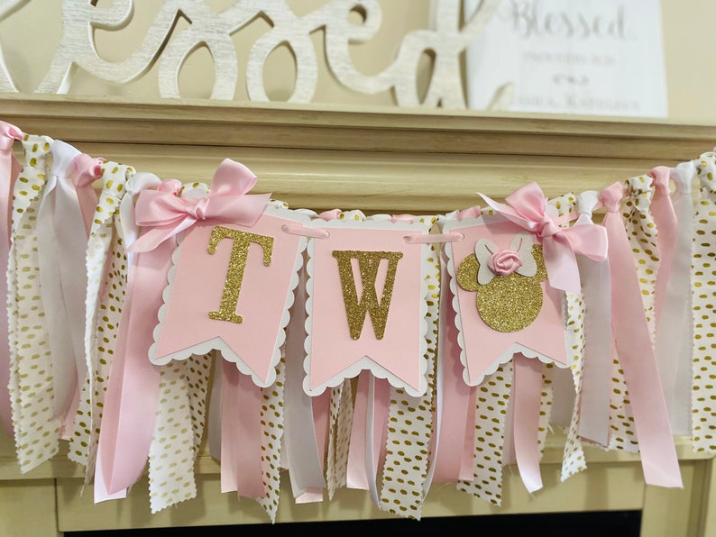 May include: A pink and gold banner with the letters "TW" and a Minnie Mouse silhouette. The banner is decorated with pink ribbons, white ribbons with gold polka dots, and gold glitter accents. A decorative piece that says "Blessed" is in the background.