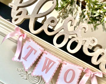 May include: Pink and white banner with the word "TWO" in pink letters. The banner is decorated with pink ribbon bows and is hanging from a wooden sign that says "blessed".